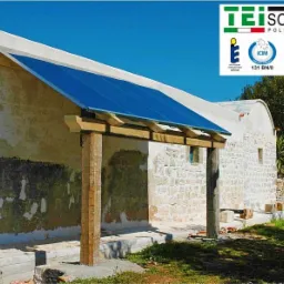 Niebieski panel słoneczny zamontowany na drewnianej konstrukcji przy budynku z kamienia, logo TEI SOLAR w prawym g&oacute;rnym rogu.