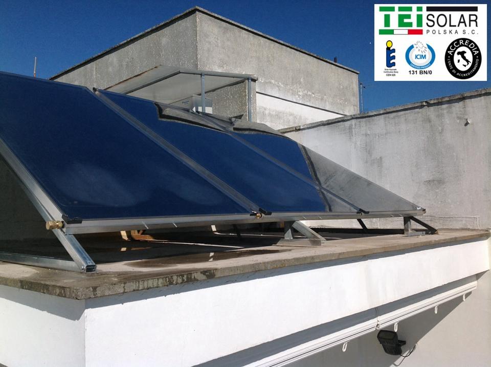 Instalacja solarna TEI Solar na płaskim dachu budynku, widoczne panele z metalową konstrukcją wsporczą, logo firmy w prawym górnym rogu, budynek w tle.