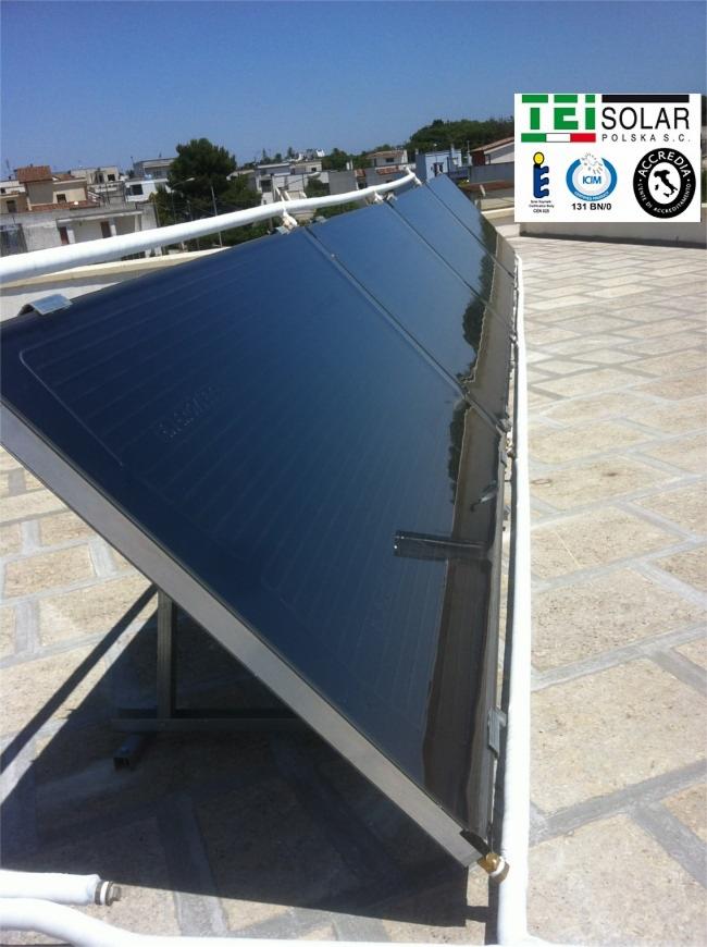 Rząd paneli słonecznych TEI Solar na dachu budynku z widocznymi budynkami w tle i niebieskim niebem.