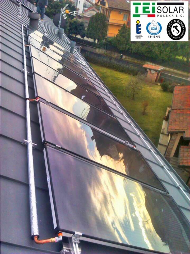 Rząd paneli słonecznych zamontowanych na dachu z szarej dachówki, odbijających błękitne niebo i chmury. Widoczne rury doprowadzające i logo firmy TEI Solar Polska S.C.