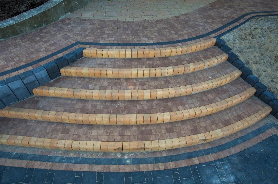 Schody z kostki brukowej w odcieniach brązu, beżu i grafitu, układające się w łukowaty wzór. Estetyczne wykończenie terenu z dbałością o detale.