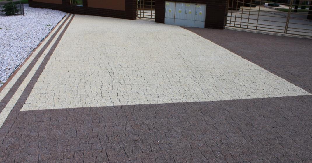 Ułożona, dwukolorowa kostka betonowa tworząca wzór na podjeździe. Widoczne ogrodzenie metalowe w tle i kamień ozdobny po lewej stronie.