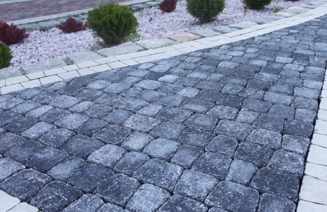 Ułożona szara kostka betonowa w jodełkę, obrzeża z jasnej kostki. W tle krzewy i jasny żwir. Estetyczne wykończenie ścieżki w ogrodzie.