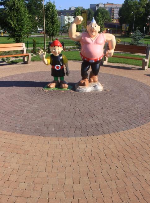 Figurki Bolka i Lolka na okrągłej nawierzchni z kostki betonowej w parku miejskim, w tle zieleń i zabudowa miejska.