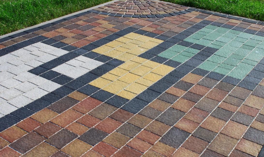 Kolorowa kostka betonowa ułożona w geometryczny wzór, z elementami białymi, żółtymi, zielonymi i brązowymi. Widoczna trawa w tle. Różnorodność kolorów i wzorów.