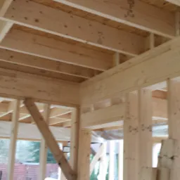 Wnętrze domu w trakcie budowy, widoczne drewniane elementy konstrukcyjne ścian i stropu, z zewnątrz widok na drzewa.