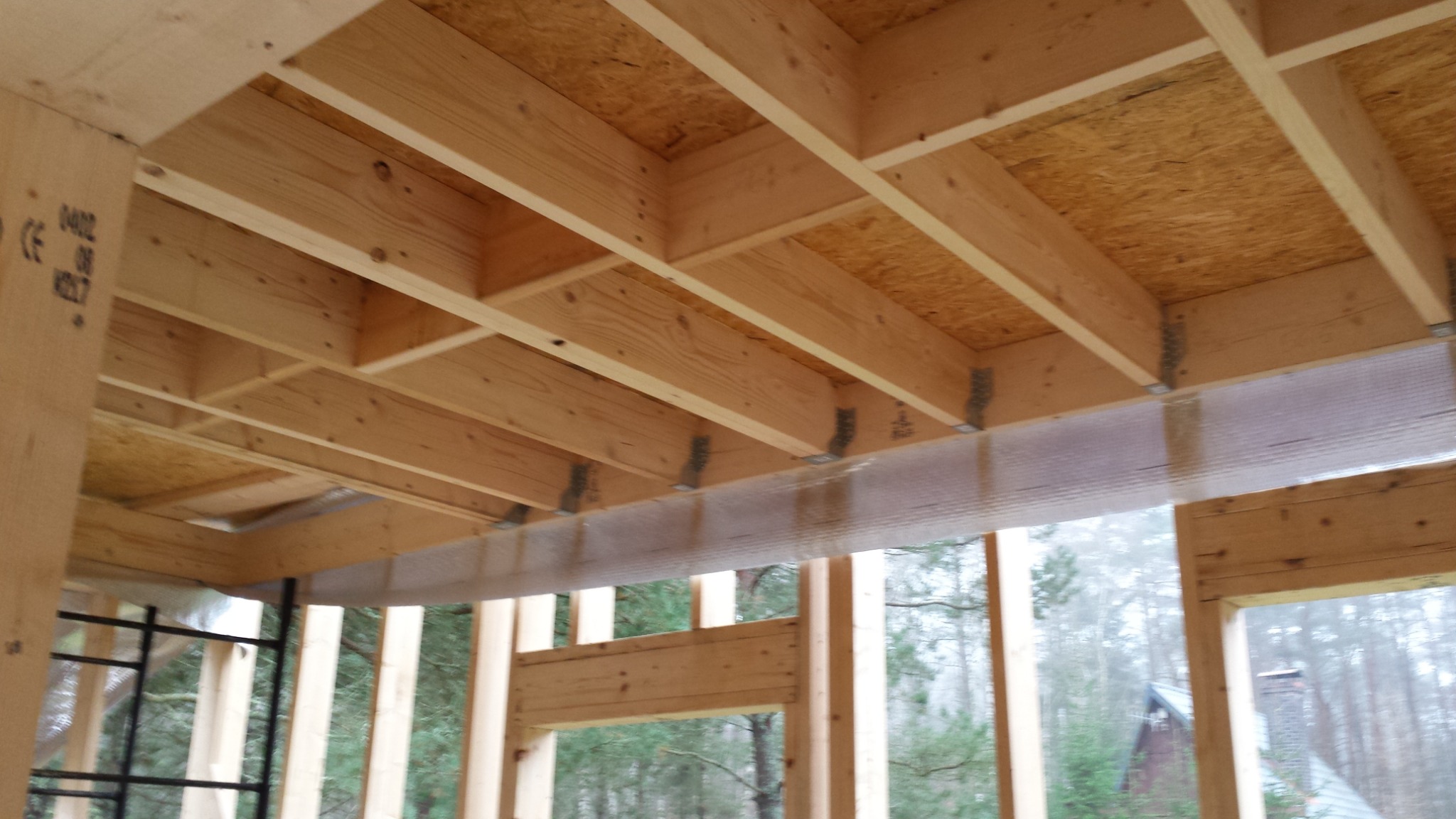 Wnętrze domu w trakcie budowy, widoczne drewniane belki stropowe, płyta OSB na suficie, folia izolacyjna i elementy konstrukcyjne ścian, z widokiem na las w tle.
