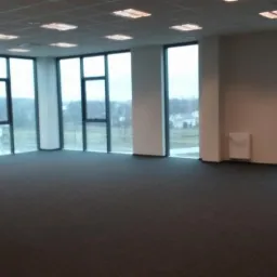 Przestronne, puste wnętrze biurowe z dużymi oknami, szarą wykładziną i białymi ścianami. Widok na zewnątrz z zielenią i budynkami. Nowoczesne oświetlenie sufitowe.