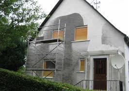 Dom w trakcie tynkowania elewacji, widoczne rusztowanie i fragmenty surowego tynku. Ujęcie z dołu, zieleń w tle.