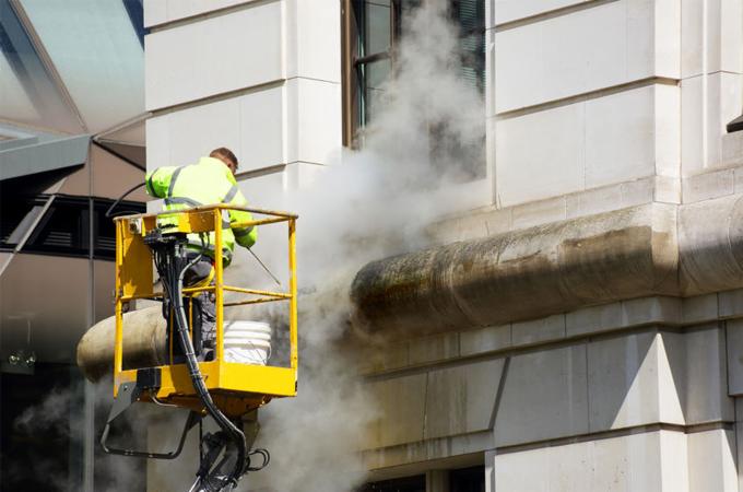 Pracownik w żółtym kombinezonie na podnośniku czyści elewację budynku strumieniem pary pod ciśnieniem, usuwając zabrudzenia z kamiennej fasady.