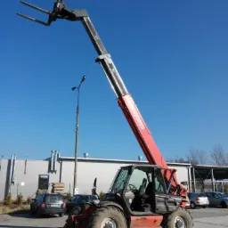 Czerwono-czarna ładowarka teleskopowa Maniscopic na placu budowy, ramię wysunięte w górę na tle błękitnego nieba i budynku z blachy falistej. W tle zaparkowane samochody.