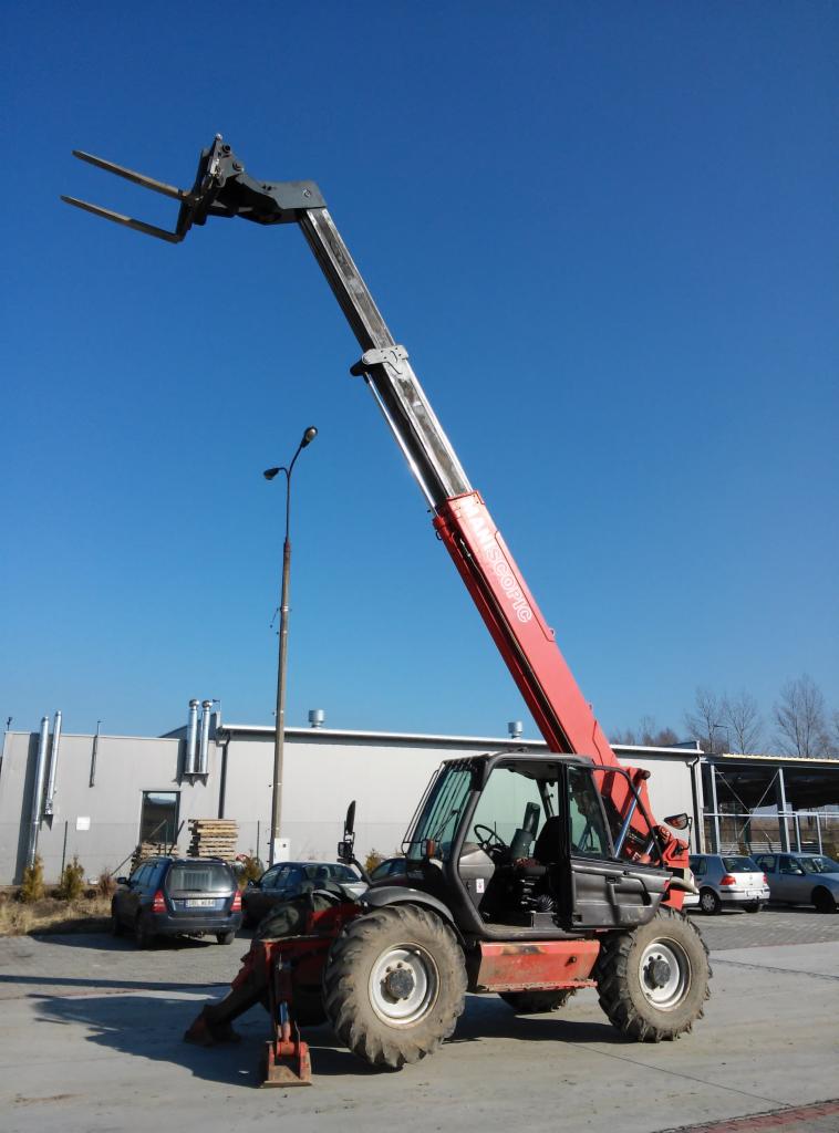 Czerwono-czarna ładowarka teleskopowa Maniscopic na placu budowy, ramię wysunięte w górę na tle błękitnego nieba i budynku z blachy falistej. W tle zaparkowane samochody.