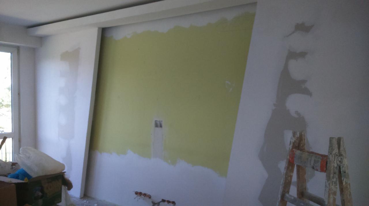 Ściana w trakcie malowania w Ostrowi Mazowieckiej. Widoczne fragmenty starej farby, drabina i narzędzia malarskie. Prace wykończeniowe w pomieszczeniu.