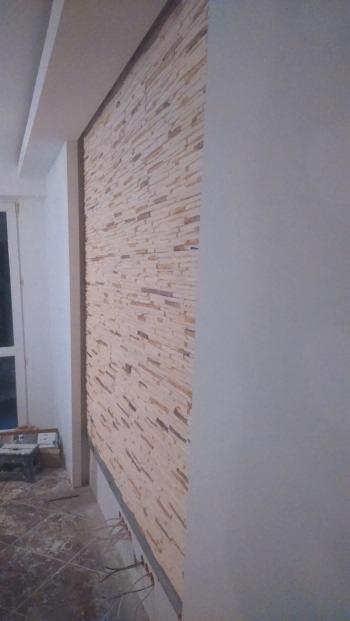 Dekoracyjna ściana z kamienia w odcieniach beżu i brązu, z widocznym fragmentem okna i sufitu. Część ściany pomalowana na szaro. Na podłodze widoczne ślady po remoncie.