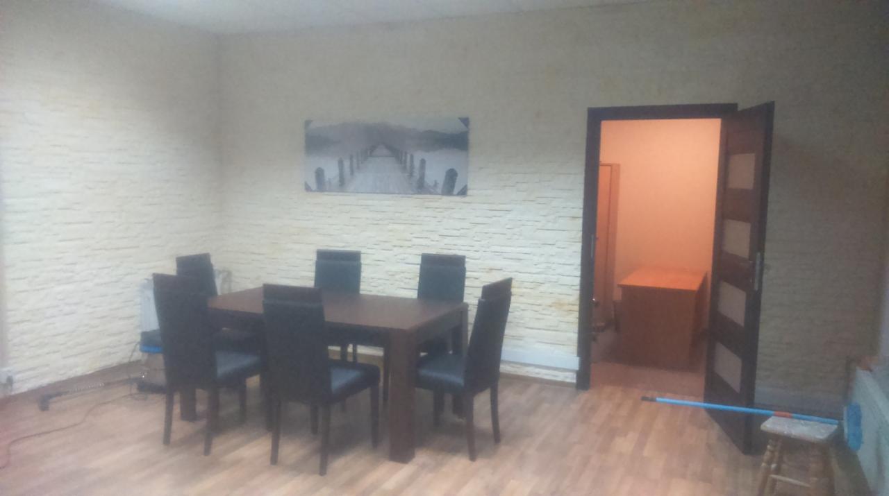 Jasne wnętrze pokoju po remoncie: teksturowana ściana, drewniana podłoga, stół z krzesłami i obraz. Otwarta, ciemna rama drzwi. Widoczne ślady po remoncie.