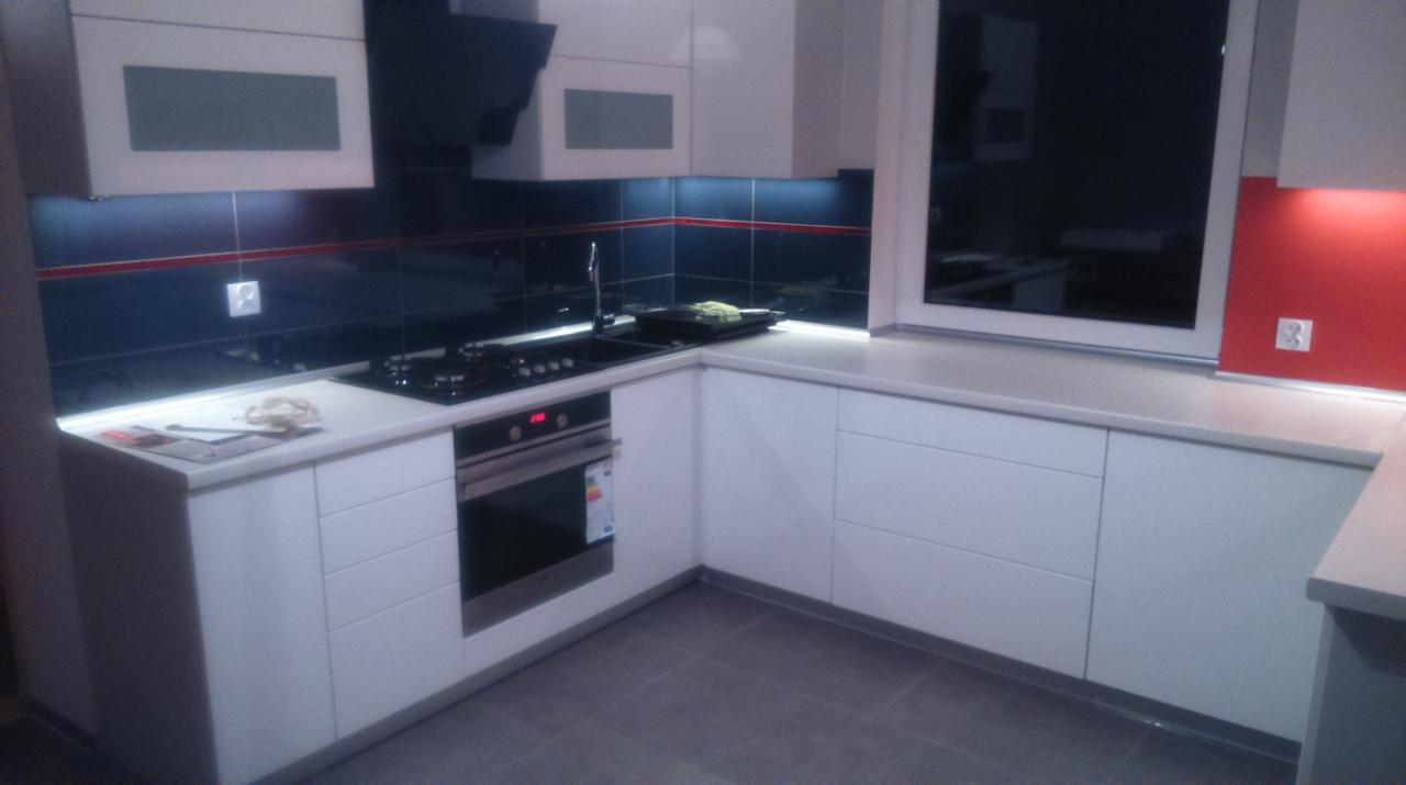 Nowoczesna kuchnia w bieli z czarnym blatem i płytkami. Oświetlenie LED podkreśla detale. Widoczny piekarnik i płyta grzewcza. Szare płytki podłogowe.