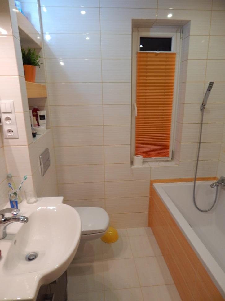 Jasna łazienka z pomarańczowymi akcentami: roleta w oknie, obudowa wanny. Umywalka z baterią, toaleta, półki z dekoracjami. Płytki na ścianach i podłodze.