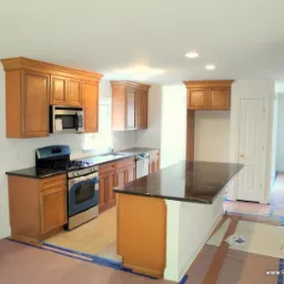 Jasna kuchnia w trakcie remontu: drewniane szafki z czarnym blatem, wyspa kuchenna z brązowym blatem, podłoga zabezpieczona folią malarską, widoczne puszki z farbą.