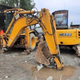 Firma szkoleniowa Zawodowy - Żółta koparko-ładowarka CAT i CASE na placu, z reklamą firmy szkoleniowej na szybie. Widoczne detale: łyżka, gąsienice, błoto. Ujęcie z bliska, podkreślające detale maszyn.