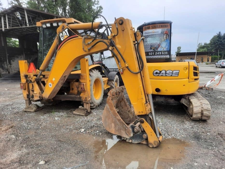 Żółta koparko-ładowarka CAT i CASE na placu, z reklamą firmy szkoleniowej na szybie. Widoczne detale: łyżka, gąsienice, błoto. Ujęcie z bliska, podkreślające detale maszyn.