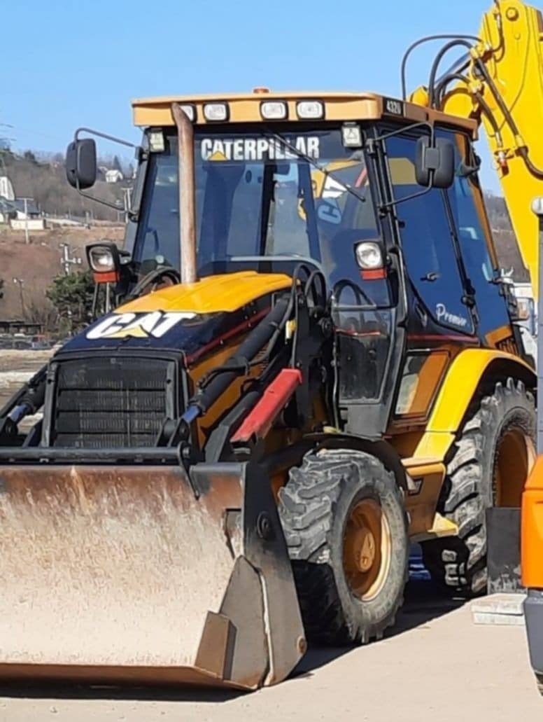 Żółto-czarna koparko-ładowarka Caterpillar z widocznym łyżką i kabiną operatora, zaparkowana na zewnątrz w słoneczny dzień. Widoczne budynki w tle.