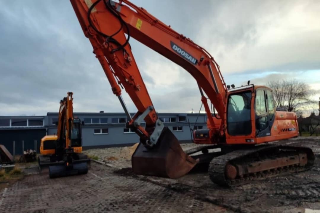 Pomarańczowa koparka Doosan na placu budowy, z drugą, mniejszą koparką w tle. Pochmurne niebo i budynek z oknami w tle. Widoczne ślady użytkowania.