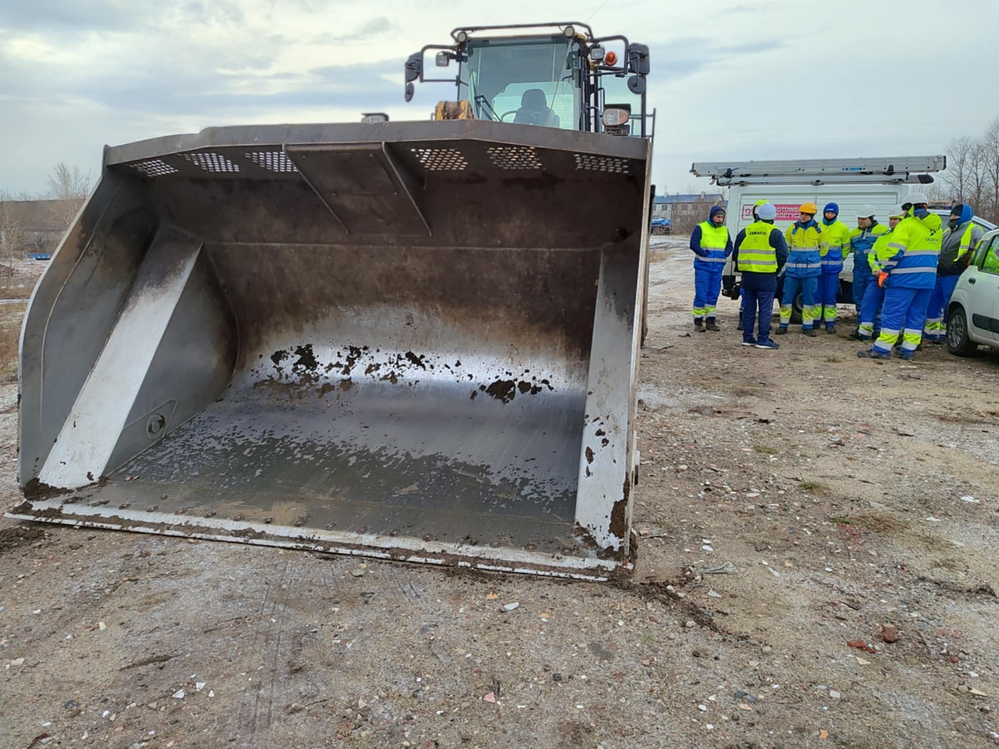 Szkolenie BHP z obsługi koparki: grupa pracowników w odblaskowych ubraniach stoi obok koparki z dużą łyżką, na placu budowy. Widok z bliska na łyżkę.