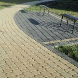 Kremowa ścieżka z kostki brukowej, obrzeża z szarej kostki. Na ścieżce dwie ławki parkowe. Widok z góry, delikatne oświetlenie.