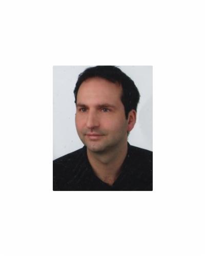 Portret mężczyzny w czarnej koszuli, o ciemnych włosach, na jasnym tle. Uśmiech lekko widoczny. Zdjęcie profilowe doradcy finansowego.