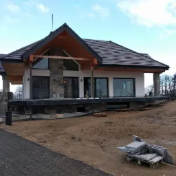 Nowoczesny dom jednorodzinny z elementami kamiennymi na elewacji, widoczny w trakcie prac wykończeniowych z ułożoną kostką brukową na podjeździe i stosem płyt chodnikowych na trawniku.