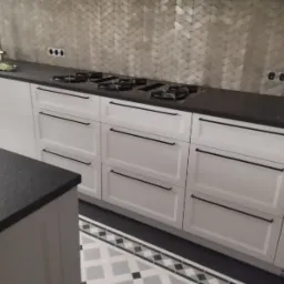 Białe szafki kuchenne z czarnymi uchwytami na tle ściany wyłożonej płytkami w jodełkę, widoczna płyta grzewcza i fragment zlewu.