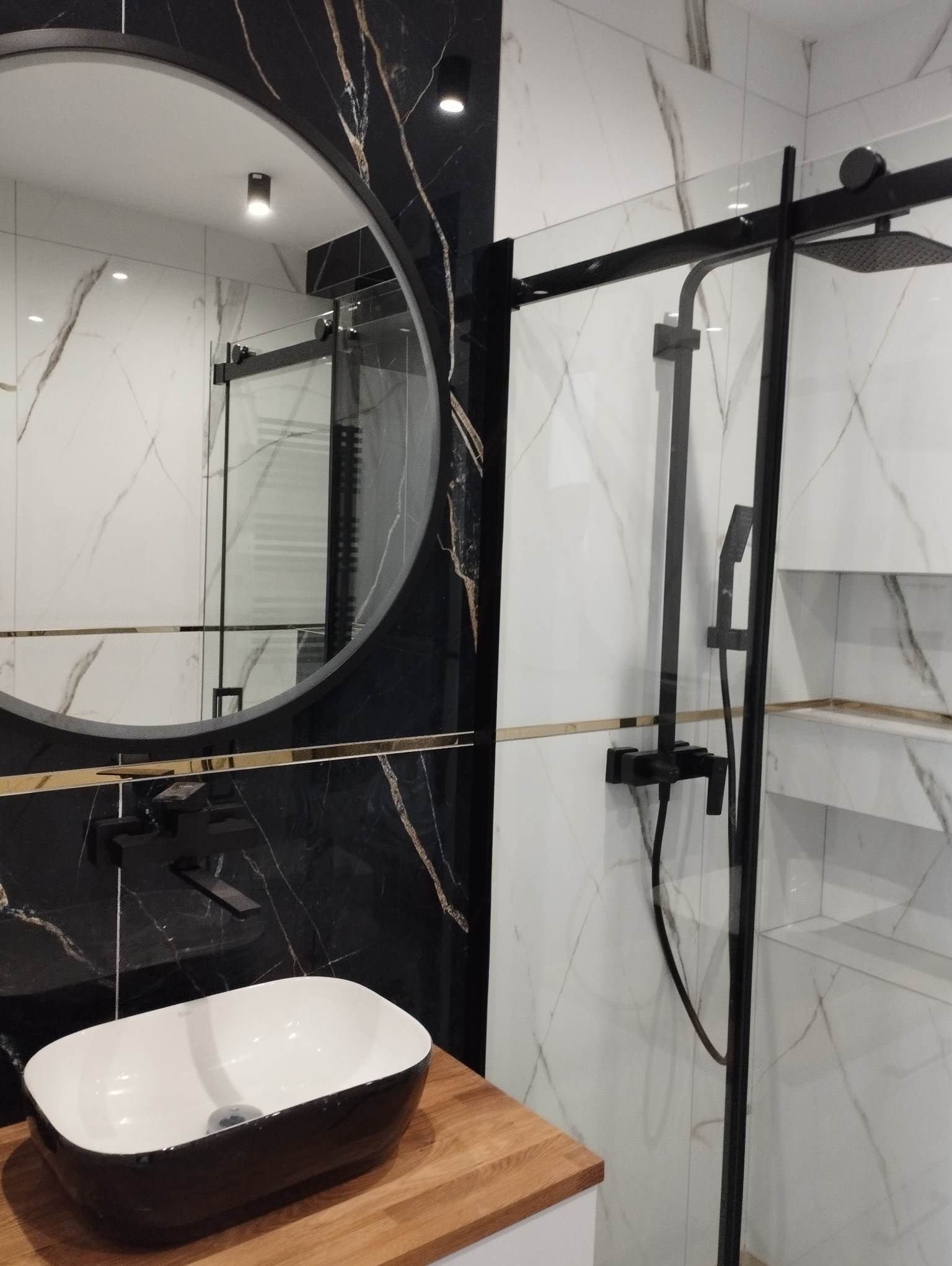 Nowoczesna łazienka z czarno-białymi marmurowymi płytkami, okrągłym lustrem z czarną ramą, drewnianym blatem pod umywalką i czarną armaturą prysznicową.