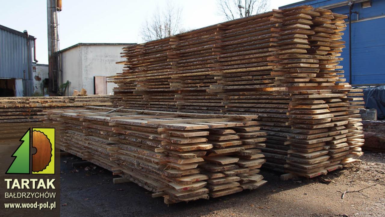 Stosy świeżo przetartej tarcicy z korą na placu składowym tartaku, z logo firmy w lewym dolnym rogu. Widoczne budynki przemysłowe w tle.