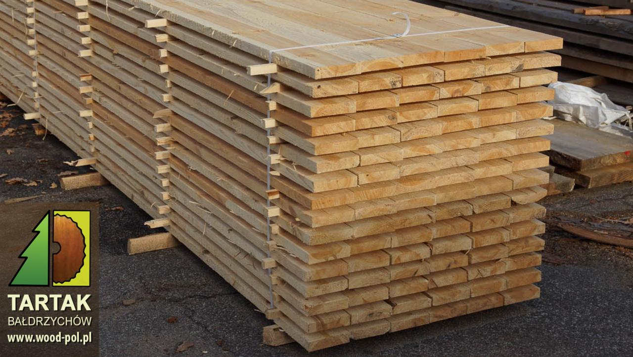 Stos tarcicy budowlanej z jasnego drewna, ułożonej warstwami na przekładkach, widoczny logo tartaku w lewym dolnym rogu.