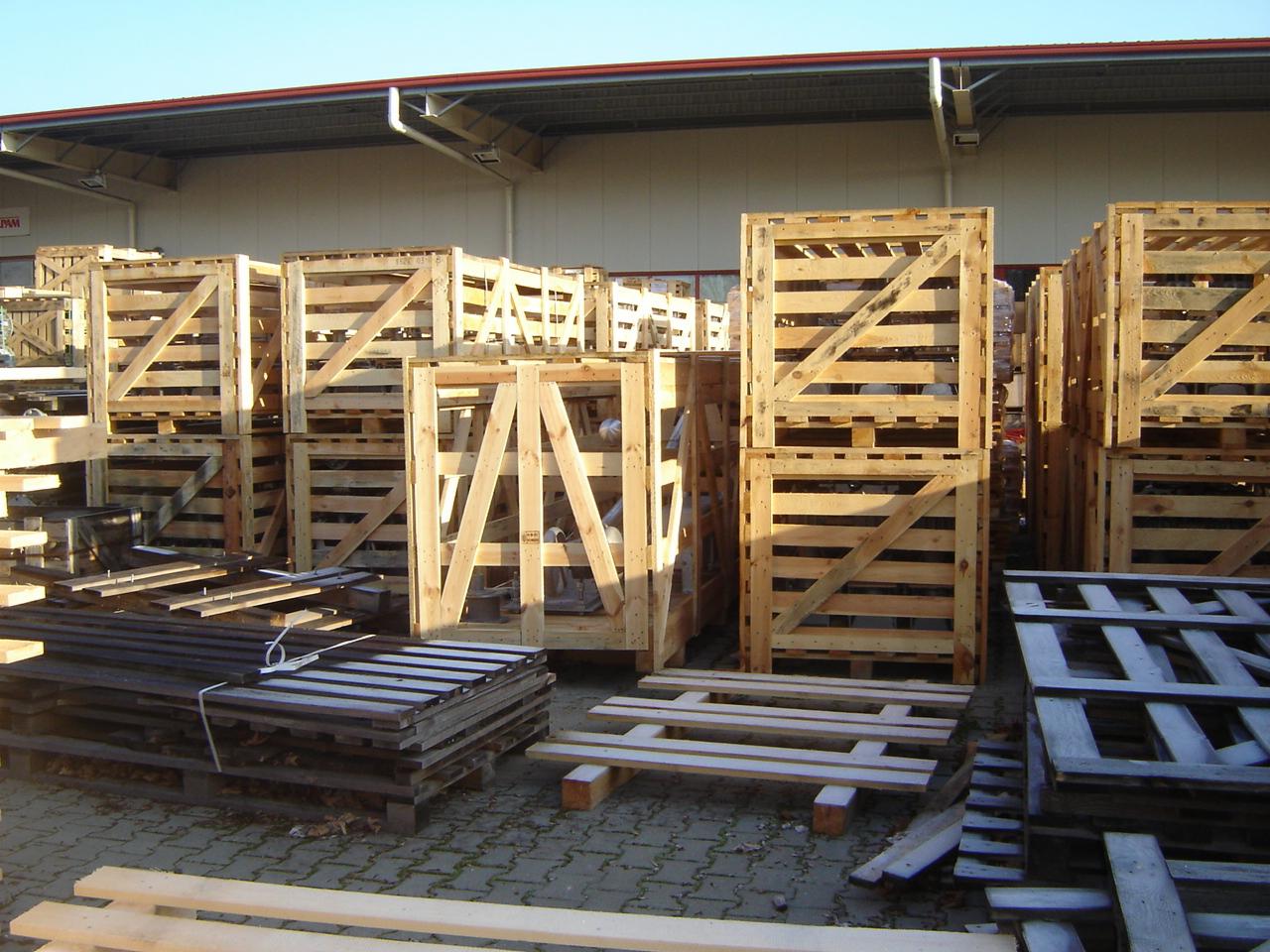 Stosy drewnianych skrzyń i palet na placu składowym, oświetlone popołudniowym słońcem, z budynkiem magazynowym w tle. Widoczne różne rodzaje i rozmiary palet.