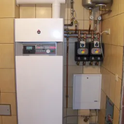 Biały, nowoczesny piec gazowy ACV HeatMaster 25TC z widocznymi dwoma zbiornikami wyrównawczymi i instalacją hydrauliczną z miedzianych rur w kotłowni wyłożonej płytkami ceramicznymi.