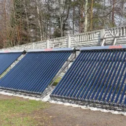 Trzy panele solarne próżniowe marki Ulrich zamontowane na betonowym fundamencie przy ogrodzeniu z prefabrykatów betonowych, na tle drzew bez liści i budynku.