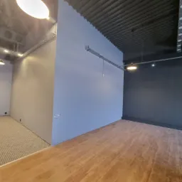 Surowe wnętrze w Wołominie przed wykończeniem: szara ściana z widoczną instalacją elektryczną, drewnopodobna podłoga i betonowy sufit z oświetleniem.