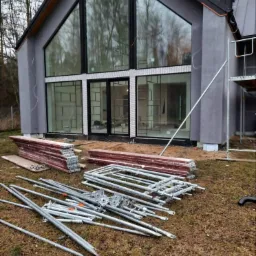 Surowy tynk na elewacji domu z dużymi oknami, widoczne rusztowania i materiały budowlane na trawie.