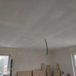 Świeżo nałożony, surowy tynk na suficie w pomieszczeniu z dwoma oknami, widoczne niedoskonałości i nierówności, przygotowany do dalszych prac wykończeniowych, z wystającymi kablami i fragmentami...