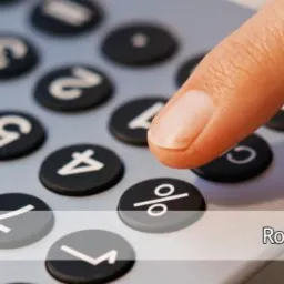 Palec naciska przycisk procent na srebrnym kalkulatorze. Rozliczenia z ZUS w tle. Detaliczny widok klawiatury kalkulatora.