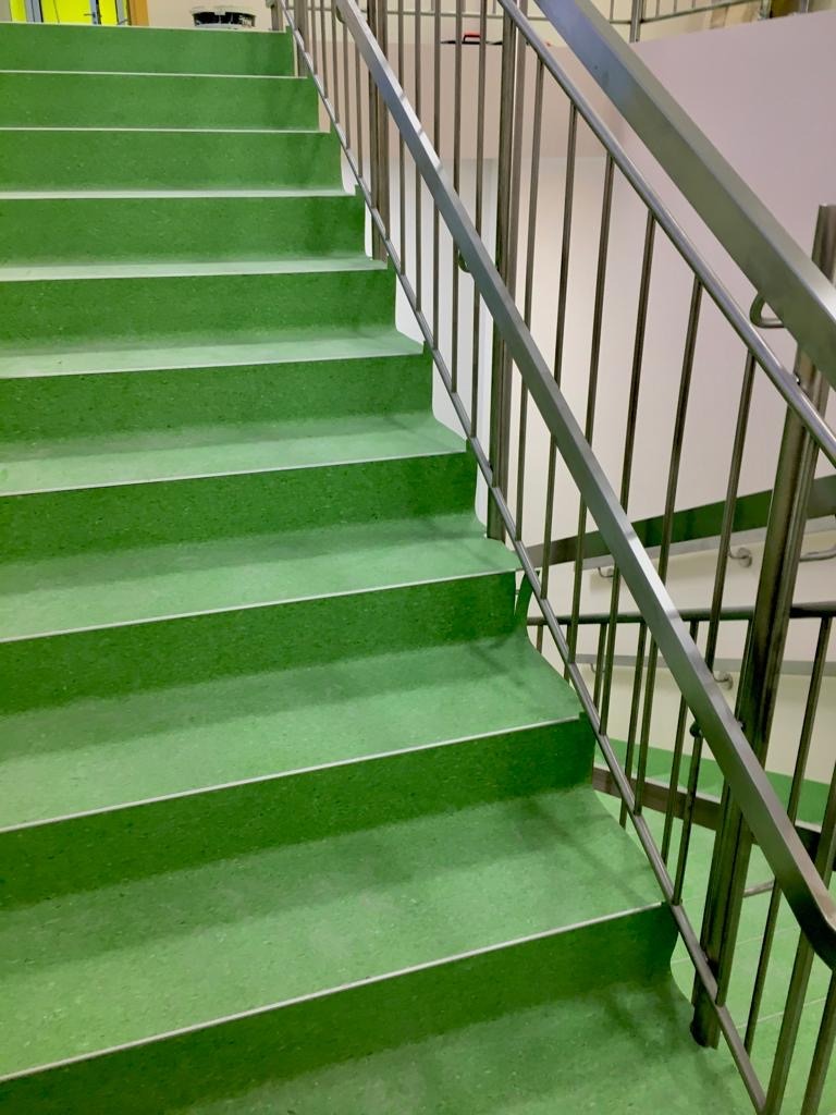 Schody pokryte zieloną, jednolitą nawierzchnią z metalową balustradą. Widok z dołu, ukazujący kilka stopni i fragment ściany w tle.