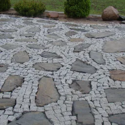 Ułożona kostka granitowa z nieregularnymi płytami kamiennymi w ogrodzie, z widocznymi krzewami i kamieniami w tle.