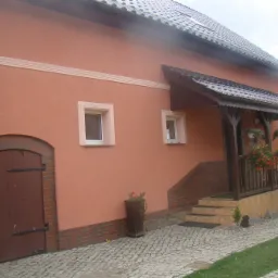 Elewacja domu jednorodzinnego w Nowym Tomyślu, pomalowana na ciepły, brzoskwiniowy odcień. Widoczne detale architektoniczne: ceglana podmurówka, okna z białymi ramami i drewniany ganek.