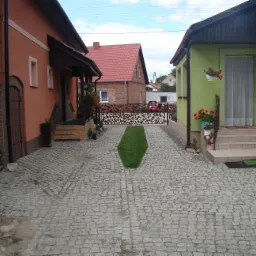 Ułożona kostka granitowa tworzy ścieżkę między dwoma budynkami o różnej kolorystyce elewacji. W tle widoczny kolejny budynek z czerwonym dachem i zaparkowane samochody.