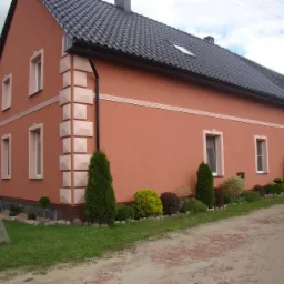 Ocieplony dom z elewacją w odcieniach brzoskwini z dekoracyjnymi elementami architektonicznymi i ciemną dachówką, otoczony zielenią i krzewami.