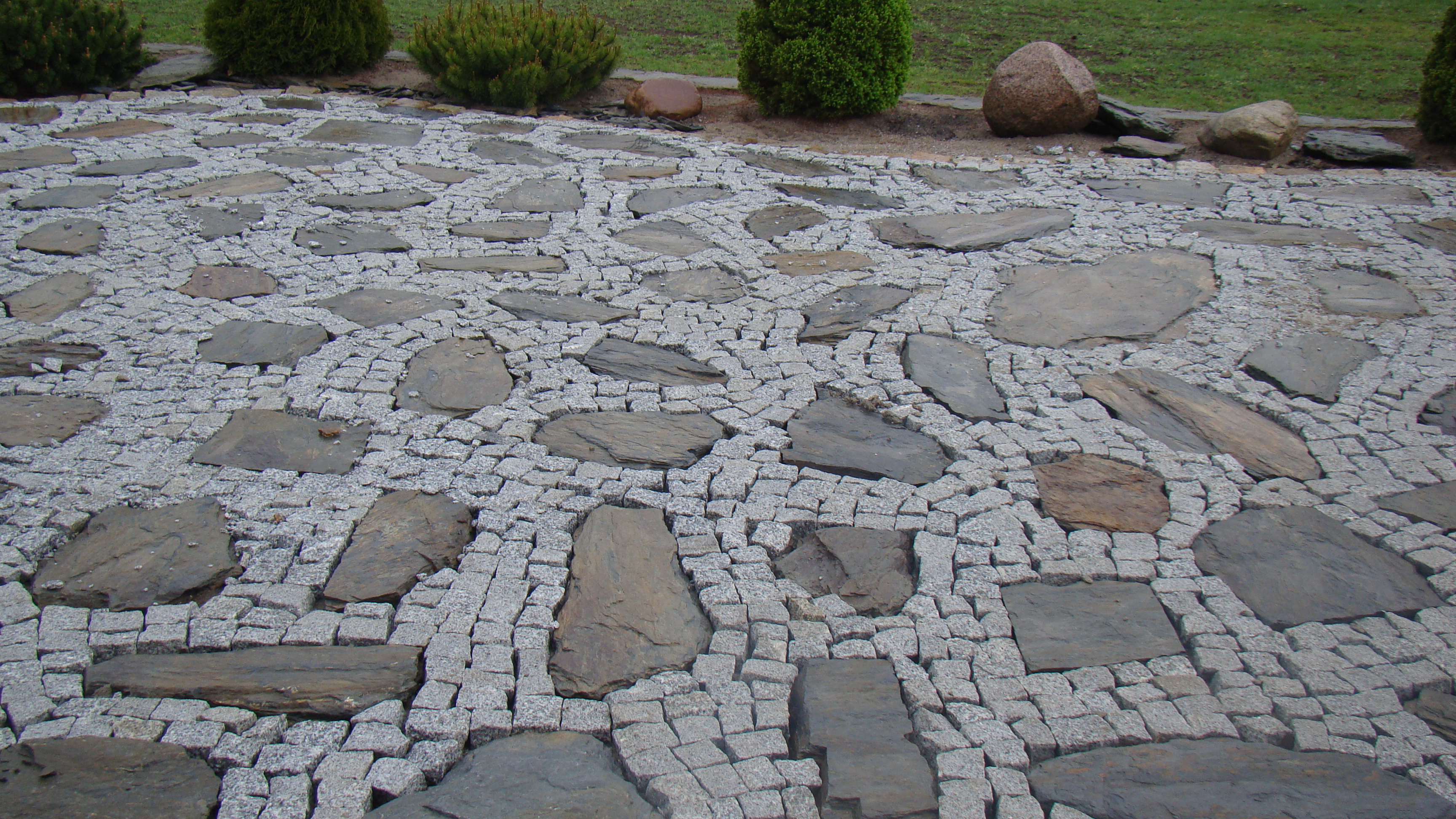 Ułożona kostka granitowa z nieregularnymi płytami kamiennymi w ogrodzie, z widocznymi krzewami i kamieniami w tle.