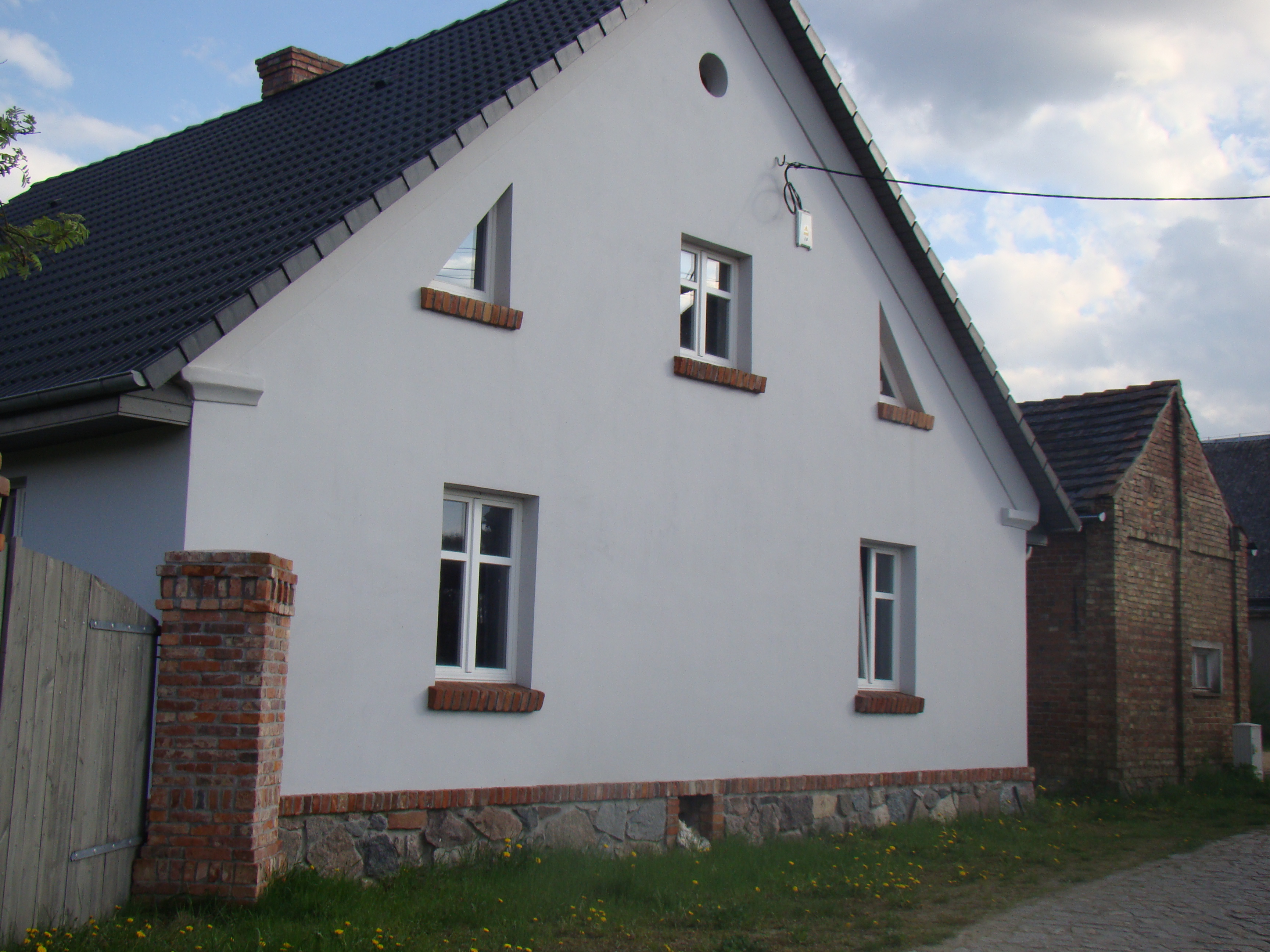 Biały dom z ciemnym dachem i ceglanymi detalami okiennymi oraz kamienno-ceglanym fundamentem. Widoczne okna o nietypowych kształtach, brama i budynek obok.