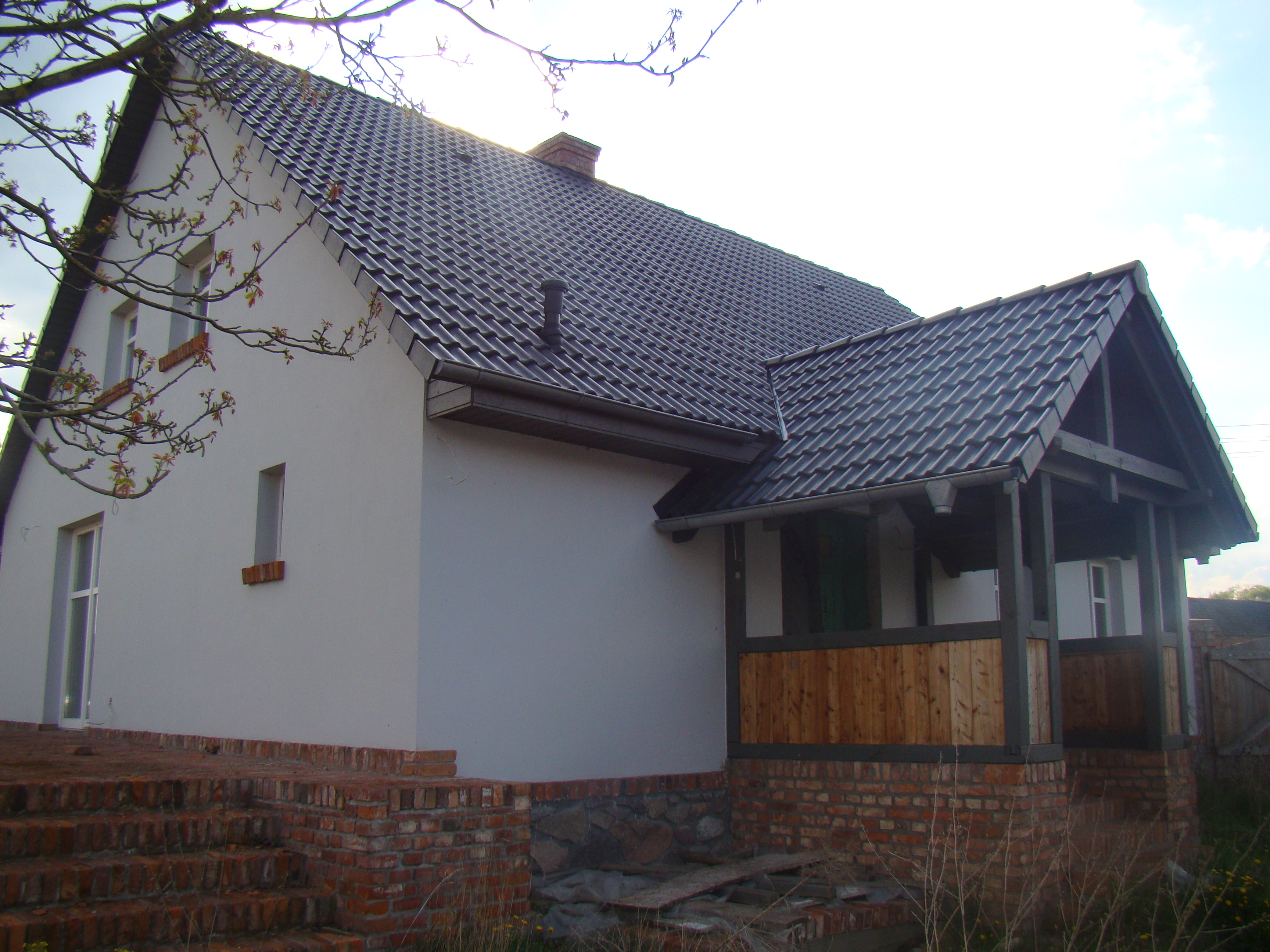 Biały dom z ciemną dachówką i ceglanymi elementami, w tym schodami. Widoczny ganek z drewnianą balustradą. Ujęcie z dołu, w tle błękitne niebo i gałęzie drzew.
