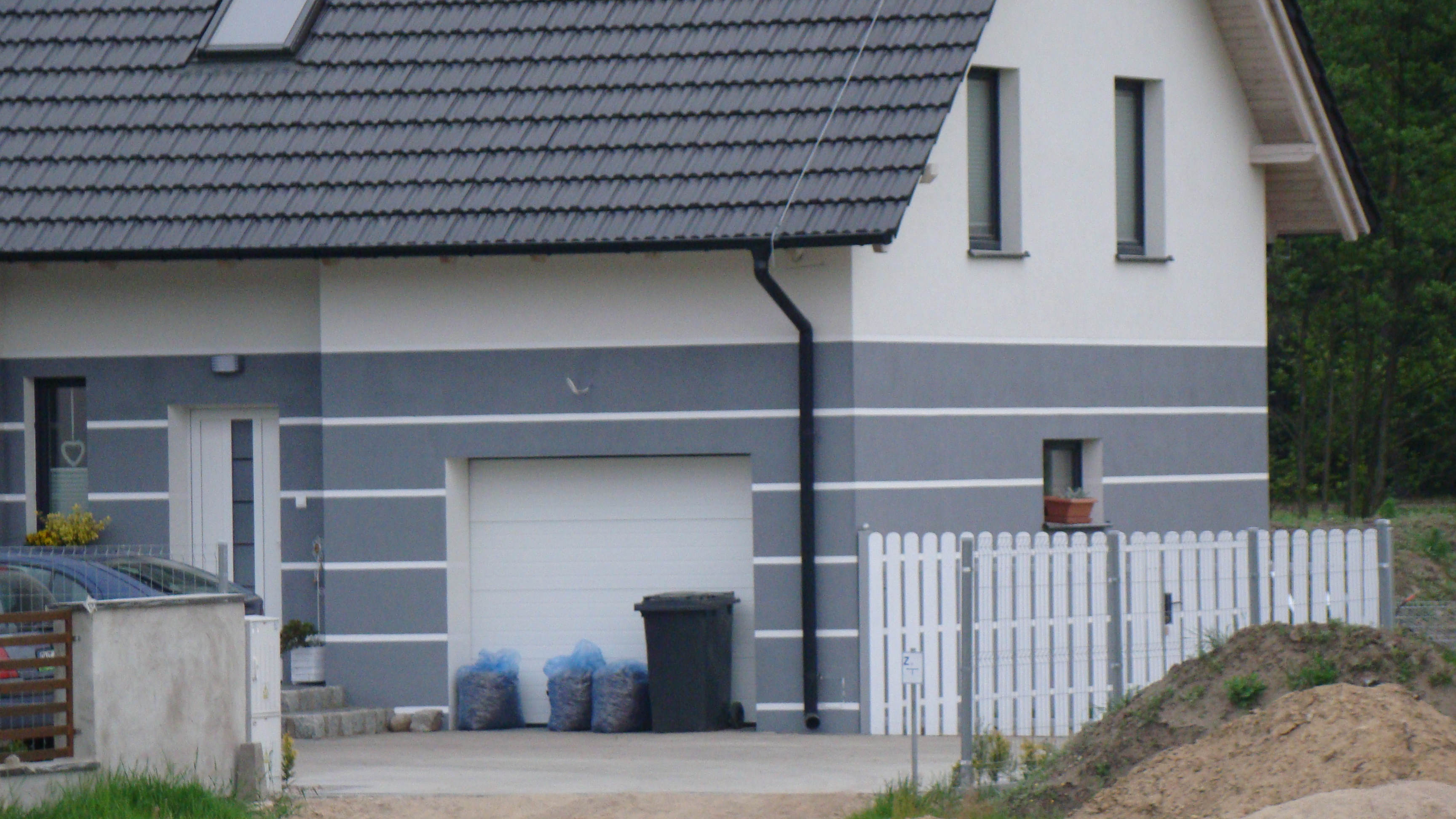 Dwukondygnacyjny dom z szarą elewacją, ciemnoszarym dachem i białymi akcentami. Widoczny garaż, brama, okna i fragment ogrodzenia. Na podjeździe worki z materiałem.